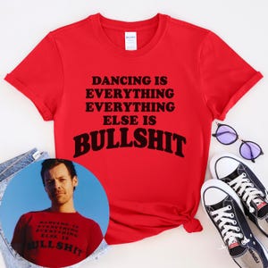 May include: A bright red short-sleeved t-shirt featuring the black text DANCING IS EVERYTHING EVERYTHING ELSE IS BULLSHIT. The shirt is displayed alongside a pair of black and white canvas sneakers and purple-tinted sunglasses, with a circular inset photo.