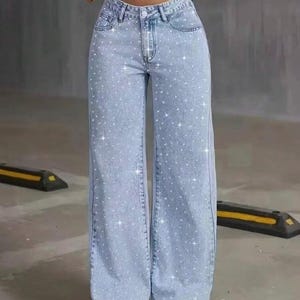 May include: Light blue denim jeans with a wide leg design. The jeans are embellished with sparkling rhinestones scattered across the fabric. The jeans have a classic five-pocket design and a button and zipper closure.