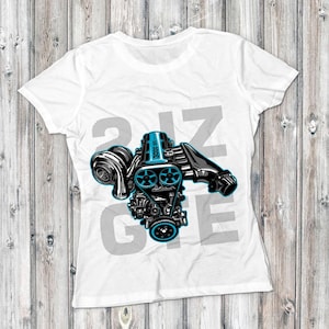 May include: White t-shirt featuring a detailed illustration of a car engine in black and teal. The design includes the text "2JZ GTE" in gray, with the engine components prominently displayed. The shirt is on a wooden background.