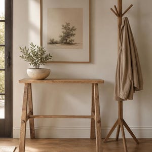 Op de afbeelding: Een rechthoekige houten sidetable met vier schuine poten en een horizontale steunbalk, een gespikkelde keramische kom met een kleine groene plant, en een houten kapstok met een beige jas, naast een ingelijste print van een boom.