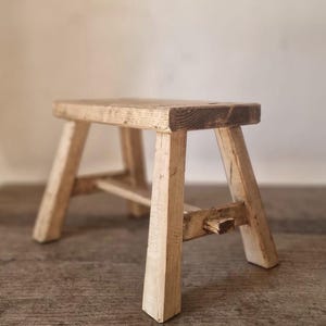 以下が含まれることがあります： 明るい茶色の風化した仕上げが施された、小さな長方形の木製スツール。このスツールは、側面にある水平なサポートストレッチャーで接続された4本の傾斜した脚が特徴で、目に見える木目とシンプルな構造を示しています。