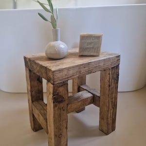 Puede incluir: Un pequeño taburete de madera rústico con aspecto envejecido. Un pequeño jarrón blanco contiene una ramita de vegetación, y una pastilla de jabón con texto está encima. El taburete está hecho de tablones ásperos.