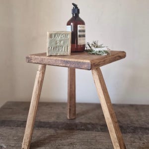May include: A three-legged wooden stool with a square top, featuring a block of Savon de Marseille soap, a brown pump bottle with an Aesop label, and a small bundle of dried herbs placed on the surface of the stool for display and utility.