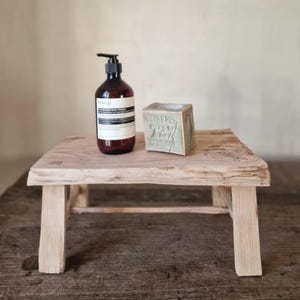 May include: A dark amber plastic pump bottle of Aesop body cleanser with a white label and a light green cube-shaped soap bar embossed with the text Extra Pur 72% d'huile, both resting on a small, rectangular, light-colored wooden stool.