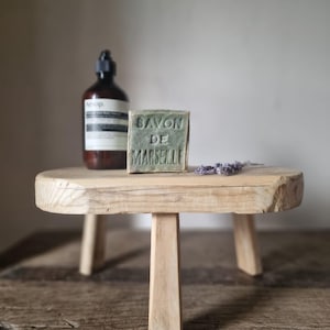 May include: A small, pale wooden stool with three legs holds a bar of green soap, a brown bottle, and a sprig of lavender. The soap is inscribed with "SAVON DE MARSEILLE". The items are displayed on a rustic wooden surface.