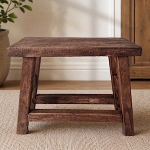 Könnte beinhalten: Ein kleiner, rechteckiger Holzhocker mit dunkelbraunem Finish. Der Hocker verfügt über vier stabile, leicht angewinkelte Beine, die durch horizontale Stützstreben an den Seiten verbunden sind, was eine stabile Basis und eine flache Sitzfläche bietet.
