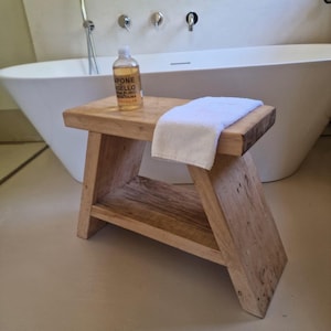 Pode incluir: Um banco retangular de madeira com uma prateleira inferior, apresentando um acabamento de grão de madeira natural castanho claro. Uma toalha branca dobrada repousa na superfície superior ao lado de uma garrafa de plástico transparente.