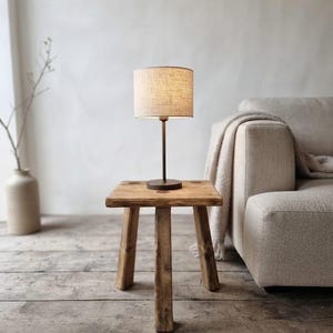 Puede incluir: Un taburete pequeño de madera de tres patas con una superficie cuadrada, que sostiene una lámpara de mesa de metal oscuro con una pantalla cilíndrica de tela beige texturizada. La lámpara está centrada y emite una luz suave y cálida.