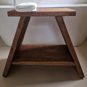 Pode incluir: Um banco de madeira castanho-escuro com um design em forma de A, apresentando um topo retangular plano e uma prateleira inferior. Uma toalha branca dobrada repousa sobre a superfície superior do banco. O veio da madeira é visível no móvel.