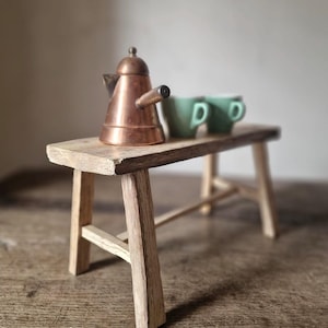 Puede incluir: Un pequeño banco de madera sostiene una cafetera de cobre y dos tazas verdes. El banco es de madera clara y tiene un aspecto rústico. La cafetera tiene un diseño clásico y las tazas son de color verde liso.