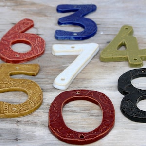 Könnte beinhalten: Ein Satz Keramik-Hausnummern in verschiedenen Farben und Stilen. Die Nummern umfassen 0, 3, 4, 5, 6, 7 und 8. Einige der Nummern haben ein Muster.