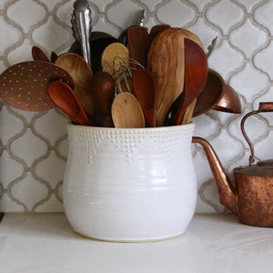 May include: A white ceramic utensil holder with a beaded rim, filled with wooden and metal kitchen utensils, sits on a white worktop next to a copper teapot with a handle.
