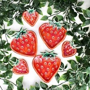 Puede incluir: Conjunto de platos de cerámica en forma de corazón diseñados para parecerse a fresas. Los platos son rojos con pequeños puntos espaciados uniformemente y tallos verdes. El arreglo está rodeado de vides de hojas verdes, creando una estética veraniega.