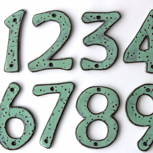 Pode incluir: Dez números de casa de cerâmica, de 1 a 10, na cor verde claro com detalhes marrons. Os números são pintados à mão com um design salpicado.
