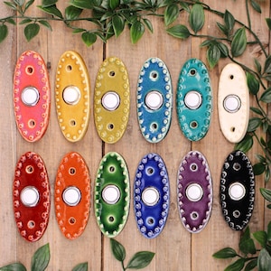 May include: Twelve oval ceramic doorbell buttons arranged in two rows, featuring a central white push button surrounded by a beaded border. The buttons come in various glossy colors including red, orange, yellow, green, blue, purple, and white.