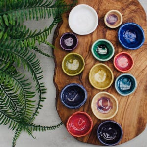 Puede incluir: Una colección de pequeños cuencos de cerámica coloridos en varios tonos de azul, verde, amarillo, morado y rojo. Los cuencos están dispuestos sobre una superficie de madera, con un plato blanco en la parte superior. Se ve un helecho en el fondo.