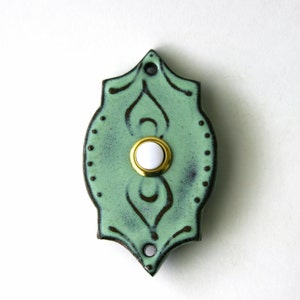 May include: A decorative ceramic doorbell button with a light green glaze and dark brown accents. The button has a white center and a gold-colored trim. The doorbell is shaped like a stylized shield with two holes at the top for mounting.