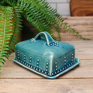 May include: A turquoise ceramic butter dish with a square shape and a handle on top. The dish features white dot detailing around the edges and on the handle. The butter dish is displayed on a wooden surface.