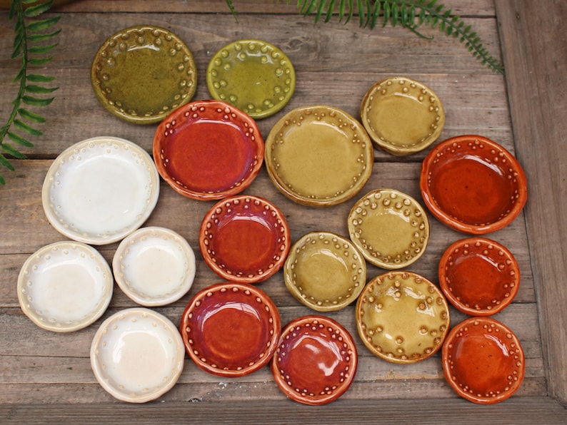 May include: A collection of small, ceramic dishes in various colors, including white, green, red, and yellow-brown. The dishes have a textured, dotted rim and are arranged on a wooden surface.