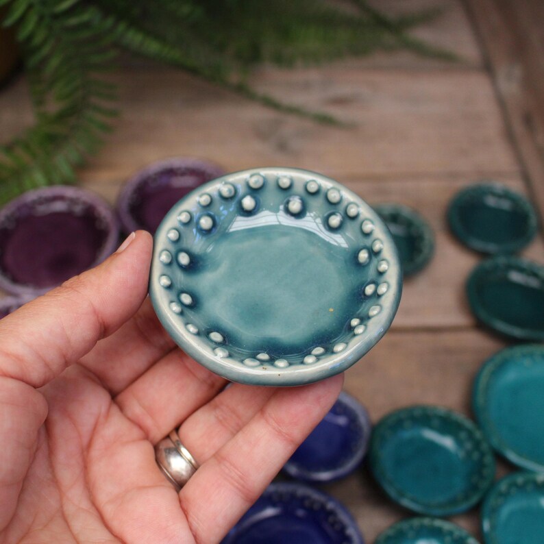 May include: A small, teal ceramic dish with a textured rim of white dots. The dish is held in a hand, with other similar dishes in various shades of blue and purple visible in the background. The dishes are on a wooden surface.