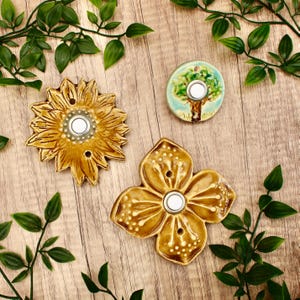 May include: Three ceramic doorbell buttons in the image. One is a sunflower, another is a four-petal flower, and the third is a round button with a tree design. All have a white button in the center. The background is a wooden surface with green leaves.