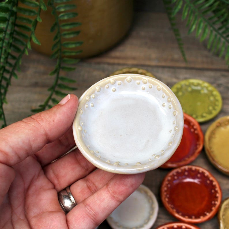 May include: A small, off-white ceramic dish with a dotted rim is held in a hand. Other colorful dishes in red, green, and yellow are visible in the background. The dish is round and appears to be handmade.