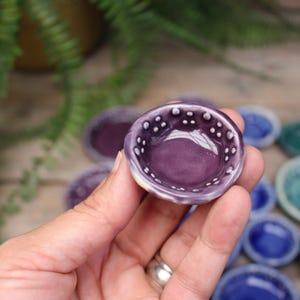 May include: A small, round, purple ceramic bowl with a white dotted rim is held in a hand. Other small bowls in shades of purple, blue, and green are in the background. The bowl is approximately 1 inch in diameter.