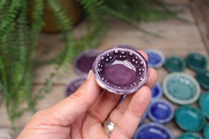 May include: A small, round, purple ceramic bowl with a white dotted rim is held in a hand. Other small bowls in shades of purple, blue, and green are in the background. The bowl is approximately 1 inch in diameter.