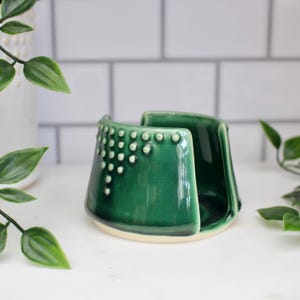K&ouml;nnte beinhalten: Ein Set aus drei Keramikbechern mit einer gesprenkelten, cremefarbenen Glasur. Jeder Becher hat ein anderes handgemaltes Design: ein blaues und gr&uuml;nes Blatt, eine gelbe Blume und eine rote Beere. Die Becher haben eine einfache, abgerundete Form.