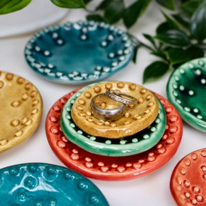 May include: A collection of small, colorful ceramic dishes with raised dot patterns. The dishes are in shades of teal, green, yellow, and red. Two rings rest on a yellow dish. The dishes are likely used for holding jewelry or small items.