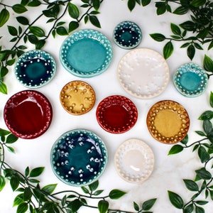 May include: A collection of small, ceramic plates in various colors, including teal, white, red, and yellow. Each plate features a dotted border and a glossy finish. The plates are arranged on a white surface with green foliage.