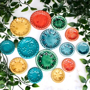 May include: A collection of colorful ceramic plates in various sizes and shades, including yellow, orange, teal, and green. Each plate features a textured, dotted rim. The plates are arranged on a white surface, with green foliage in the background.
