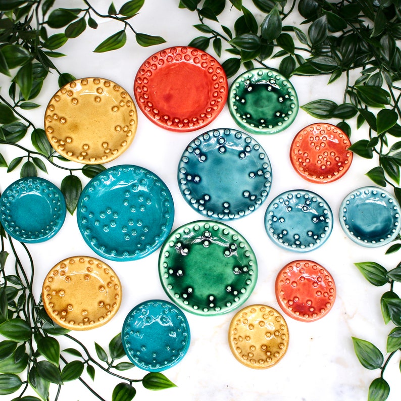 May include: A collection of colorful ceramic plates in various sizes and shades, including yellow, orange, teal, and green. Each plate features a textured, dotted rim. The plates are arranged on a white surface, with green foliage in the background.