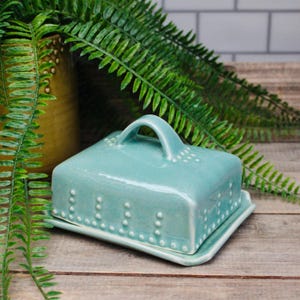 May include: A turquoise ceramic butter dish with a square lid and handle. The lid and base are decorated with small, raised white dots. The dish sits on a wooden surface, with green fern leaves in the background.