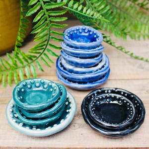 May include: A collection of handmade ceramic bowls in shades of blue, teal, and black. The bowls are stacked in varying sizes, with a textured rim and a glossy finish. The arrangement is set on a wooden surface with green foliage in the background.