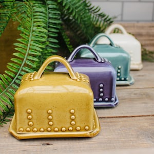 May include: A row of colorful ceramic butter dishes with handles. The front dish is mustard yellow, followed by purple, teal, and white. Each dish has a square base and is decorated with small, round studs. The dishes sit on a wooden surface.