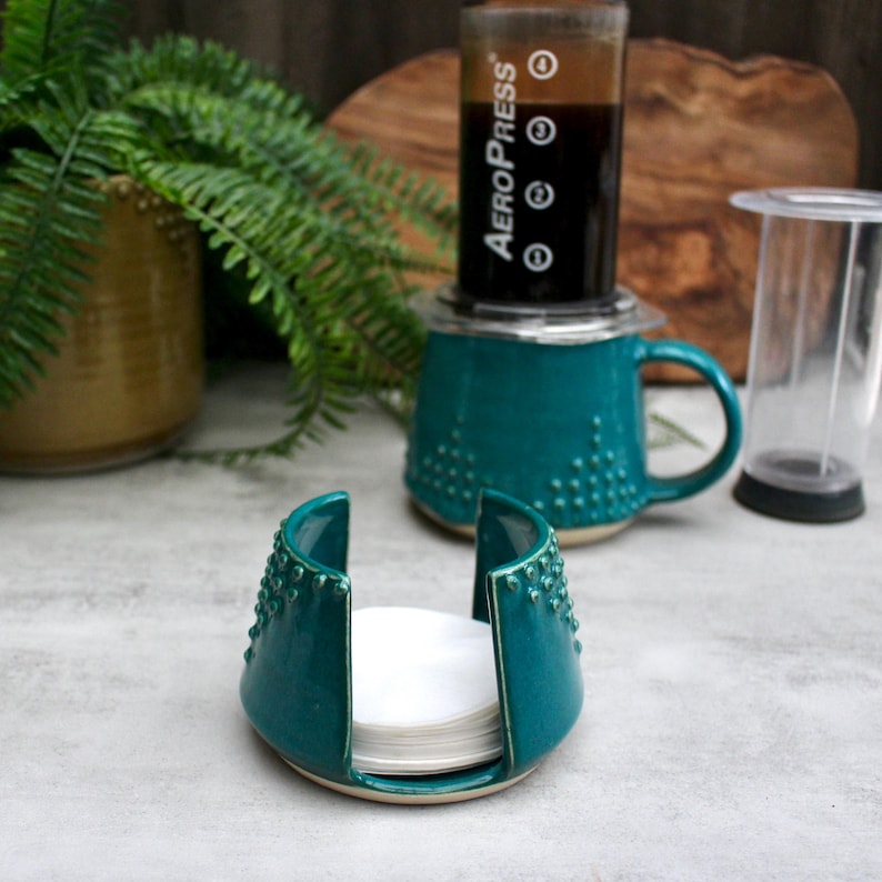 K&ouml;nnte beinhalten: T&uuml;rkisfarbener Keramik-Kaffeefilterhalter mit einem Stapel wei&szlig;er Filter. Der Halter hat ein strukturiertes, gepunktetes Design. Im Hintergrund befindet sich eine t&uuml;rkisfarbene Tasse mit einer Kaffeemaschine. Ein durchsichtiger Kunststoffbeh&auml;lter ist ebenfalls sichtbar.
