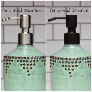 May include: Two ceramic soap dispensers with a light green glaze and brown dots. The dispensers have different colored pumps: one is brushed stainless steel and the other is brushed bronze. The text "Brushed Stainless" and "Brushed Bronze" is above each dispenser.