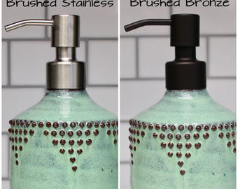 Puede incluir: Dos dispensadores de jabón de cerámica con un esmalte verde claro y puntos marrones. Los dispensadores tienen bombas de colores diferentes: uno es de acero inoxidable cepillado y el otro es de bronce cepillado. El texto "Brushed Stainless" y "Brushed Bronze" está encima de cada dispensador.