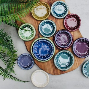 May include: A collection of small, colorful ceramic dishes with a textured rim and a central well. The dishes are in various colors, including blue, green, purple, and yellow. They are arranged on a wooden cutting board with green foliage.