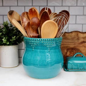 May include: A turquoise ceramic utensil holder filled with wooden spoons and a whisk. The holder has a textured rim and a rounded shape. A small butter dish and a wooden cutting board are also visible.