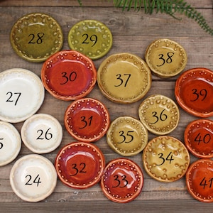 May include: A collection of small, ceramic dishes in various colors, including white, red, and yellow-green. Each dish has a unique number from 24 to 41, and a decorative dotted rim. The dishes are arranged on a wooden surface.