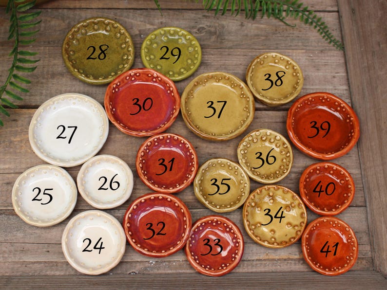 May include: A collection of small, ceramic dishes in various colors, including white, red, and yellow-green. Each dish has a unique number from 24 to 41, and a decorative dotted rim. The dishes are arranged on a wooden surface.