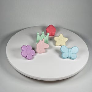 May include: A collection of colorful, decorative shapes including a flower, a unicorn, a butterfly, a star, a cupcake, and a crown. The shapes are in pastel colors: purple, pink, light blue, yellow, light green, and red. They are arranged on a white circular surface.