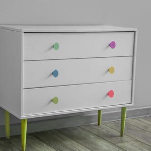 Op de afbeelding: Een witte dressoir met drie lades, elk met een gekleurde knop. De knoppen zijn groen, paars, blauw, geel en rood. De poten zijn limoengroen. De kast staat tegen een grijze muur.