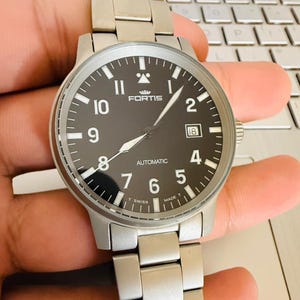 May include: A stainless steel Fortis automatic wristwatch with a black dial. The watch has white numerals, hands, and a date window at the 3 o'clock position. The watch band is made of stainless steel.