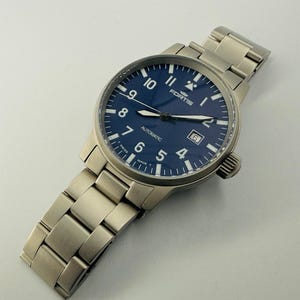 May include: A stainless steel Fortis automatic watch with a blue dial. The watch features white numerals, hands, and markers, with a date window at the 3 o'clock position. The watch has a stainless steel strap.