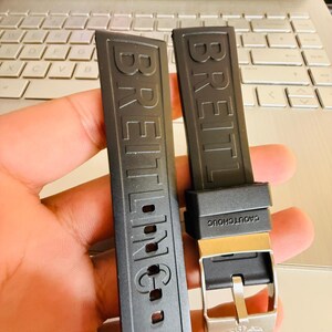 Puede incluir: Dos correas de reloj negras con el nombre de la marca "BREITLING" en relieve. Una correa tiene una hebilla plateada y el texto "CAOUTCHOUC". Las correas se muestran contra un teclado de ordenador port&aacute;til.