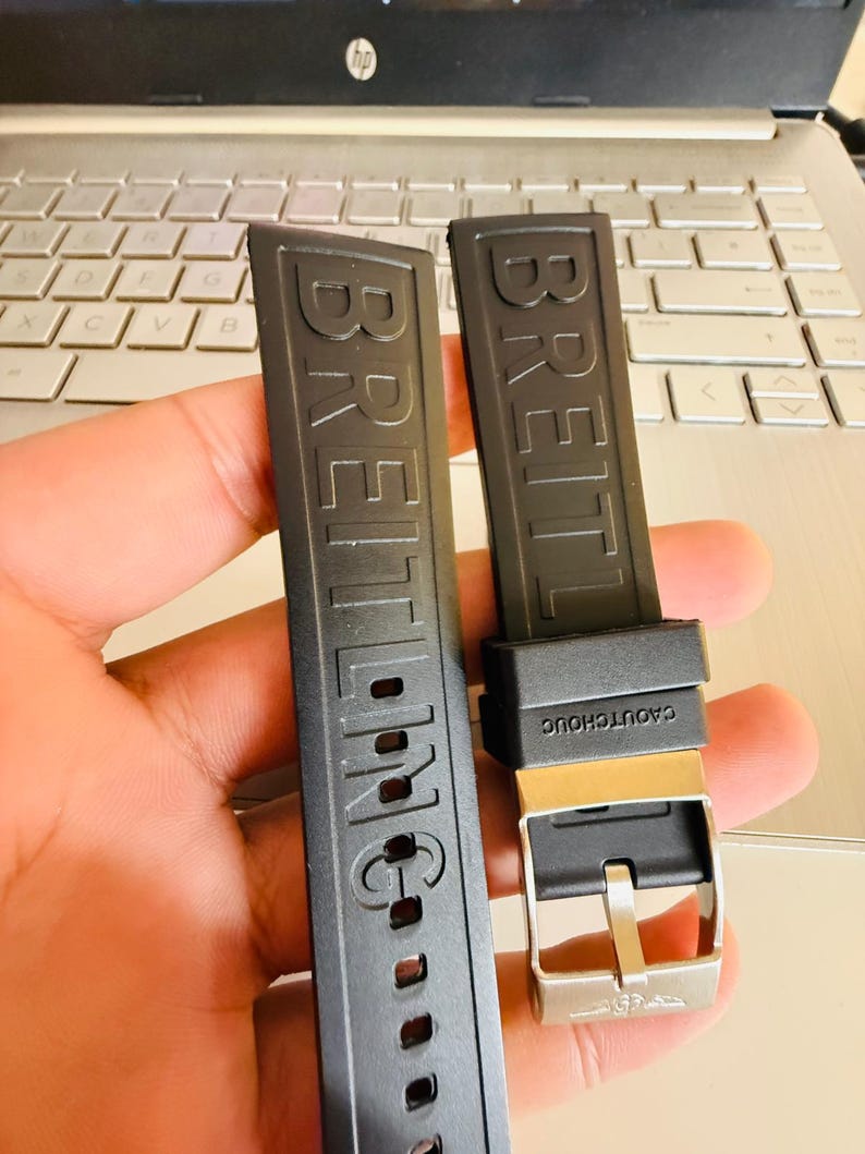 Puede incluir: Dos correas de reloj negras con el nombre de la marca "BREITLING" en relieve. Una correa tiene una hebilla plateada y el texto "CAOUTCHOUC". Las correas se muestran contra un teclado de ordenador port&aacute;til.