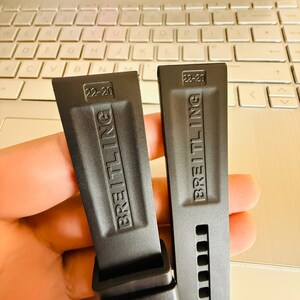 Puede incluir: Dos correas de reloj negras con el nombre de la marca "BREITLING" en relieve y la talla "22-20". Las correas se muestran frente a un teclado de ordenador port&aacute;til.